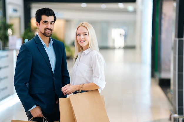 Portrait of beautiful smiling young couple holding shopping paper bags with purchases and looking at