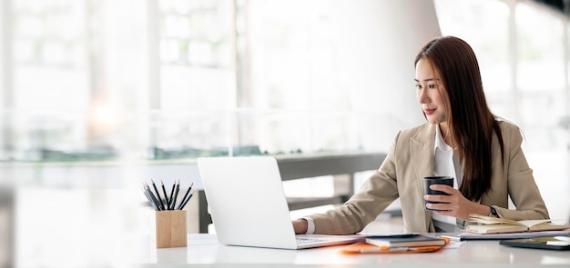 Portrait of beautiful smiling young entrepreneur businesswoman holding cup of coffee and working on laptop computer at office