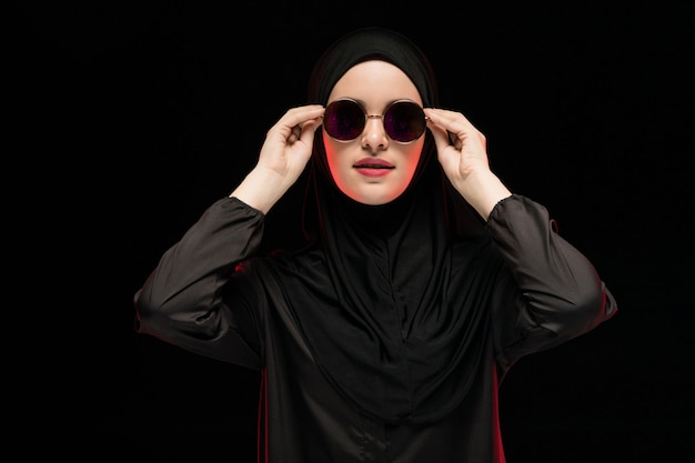 Portrait of beautiful stylish young muslim woman wearing black hijab and sunglasses as modern eastern fashion concept posing on black background