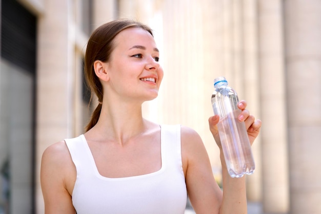 Portrait of beautiful teenager pretty girl young happy thirsty woman is drinking pure fresh water from bottle at warm sunny summer day outdoors smiling