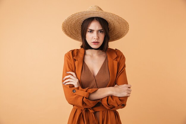 Portrait of a beautiful upset young woman wearing straw hat standing isolated over beige wall, arms folded