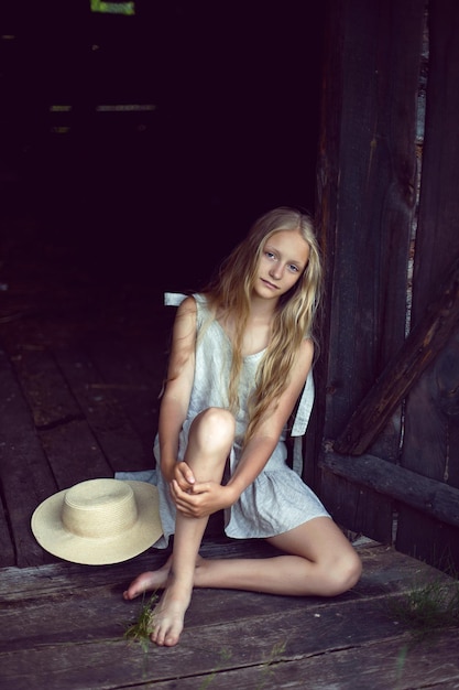 Portrait of a beautiful village girl blonde with long hair in a dress and hat sitting by a wooden old barn in summer