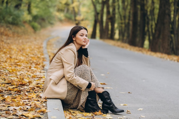 Portrait of a beautiful woman in a beige coat holding yellow maple leaves while sitting in an autumn park