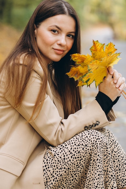 Portrait of a beautiful woman in a beige coat holding yellow maple leaves while sitting in an autumn park