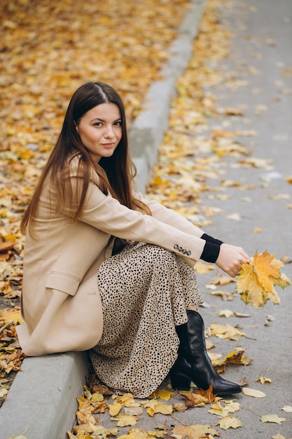 Portrait of a beautiful woman in a beige coat holding yellow maple leaves while sitting in an autumn park