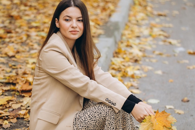 Portrait of a beautiful woman in a beige coat holding yellow maple leaves while sitting in an autumn park