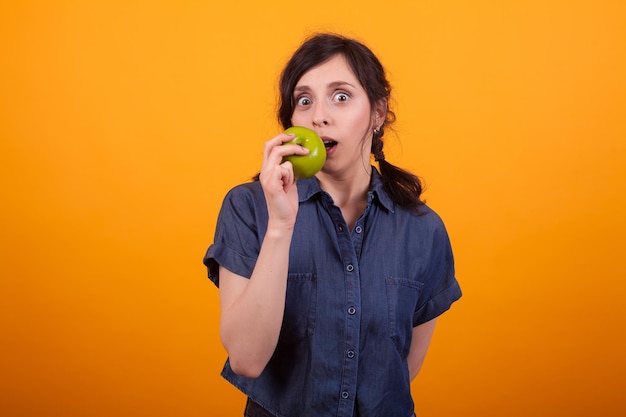 Portrait of beautiful woman bitting a green apple and looking at the camera in studio over yellow background. Young woman eating healthy fruits. Cheerful woman eating an organing apple.