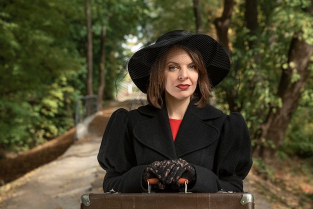 Portrait of beautiful woman in black hat with brims classic jaket and gloves hands on old suitcase Forest background