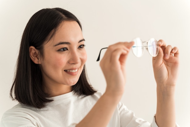 Portrait of the beautiful woman checking the cleanliness of glasses while holding it and looking on it. Girl posing on a white wall background