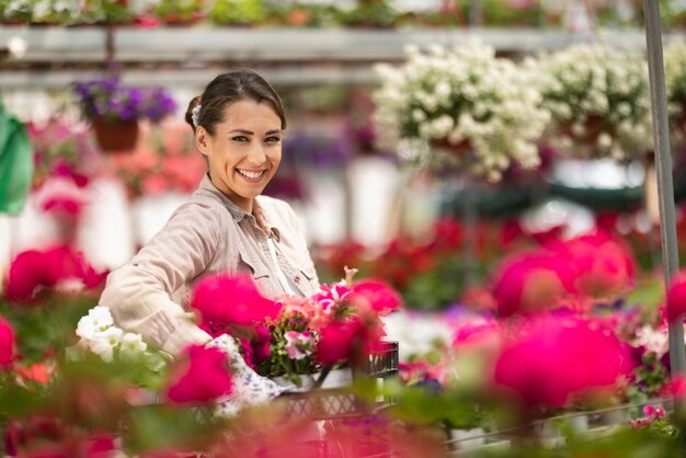 Portrait of beautiful woman florist in a greenhouse. She is holding crate with beautiful flowers and looking at camera.