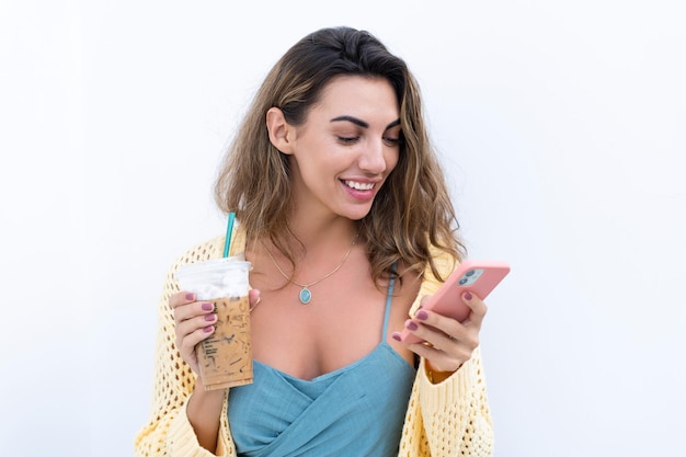 Portrait of beautiful woman in green summer dress on white background natural daylight holding iced coffee cappuccino and phone thoughtful smiling