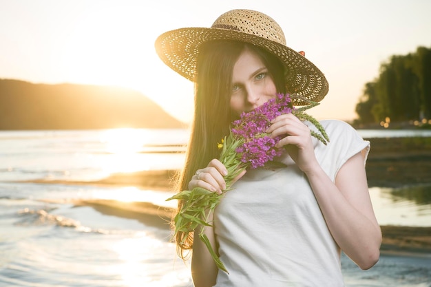 Portrait of a beautiful woman in a hat on a background of a sunset. Girl with a bouquet of flowers