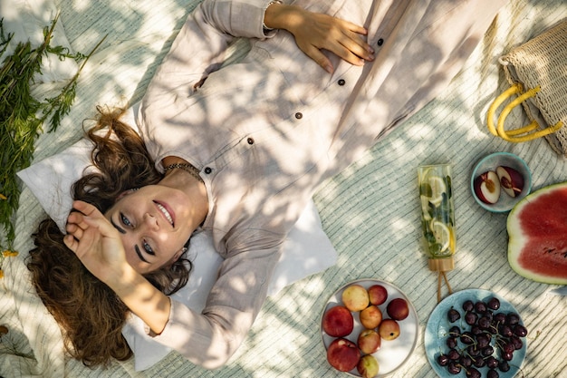 Portrait of beautiful woman posing lying on plaid at summer garden park a book fruit a bag and a wat