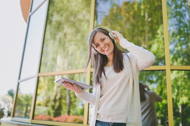 Portrait of beautiful woman wearing light casual clothes. Girl holding tablet pc computer, listening music in headphones in city park near mirror building outdoors on spring nature. Lifestyle concept.