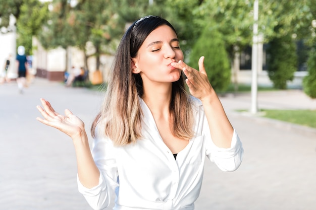 Portrait of beautiful woman in white shirt and hair bezel with mouth full of food licking her fingers outdoor in city park and enjoying junk but tasty fast food while walking