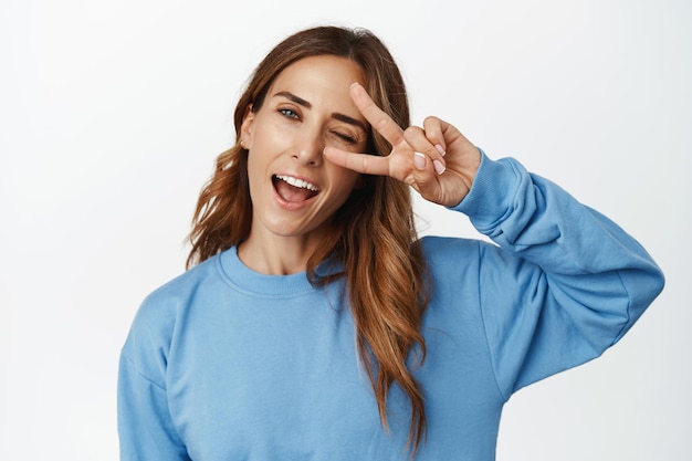 Portrait of beautiful woman winking, showing peace v-sign near eye and smiling carefree, having fun, positive emotions, standing against white background