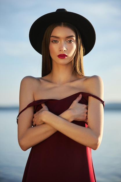 Portrait of a beautiful woman with long hair in a big round black hat. Beauty skin face, bare shoulders. Girl golden light at sunset