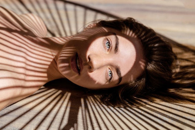 Portrait of beautiful woman with shadows of palm leaf on her face. Concept of sunbathing, vacation.