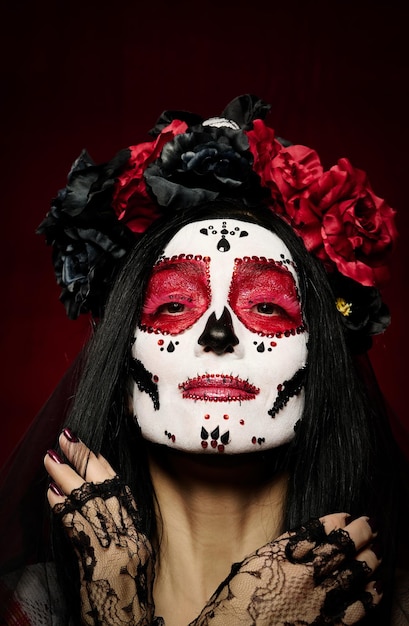 Portrait of a beautiful woman with a sugar skull makeup with a wreath of flowers on her head red background