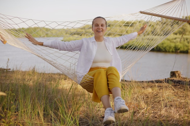 Portrait of beautiful young adult woman sitting on hammock by the water looking at camera resting spreads hands laughing happily expressing positive emotions