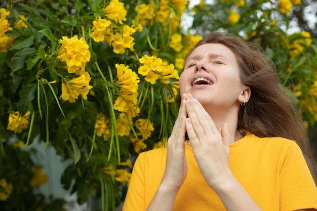 Portrait of beautiful young allergic woman is suffering from pollen allergy or cold on natural flower flowering tree background at spring or sunny summer day sneezes blowing her runny nose rubs eyes