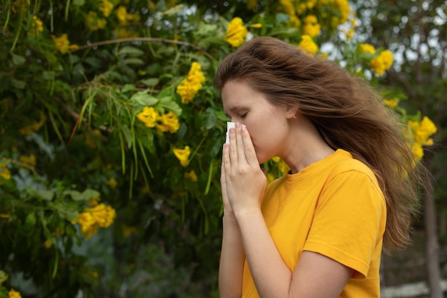 Portrait of beautiful young allergic woman is suffering from pollen allergy or cold on natural flower flowering tree background at spring or sunny summer day sneezes blowing her runny nose rubs eyes