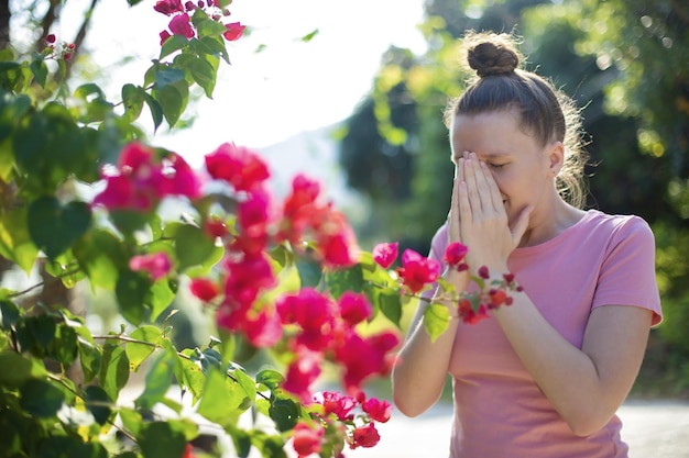 Portrait of beautiful young allergic woman is suffering from pollen allergy or cold on natural flower flowering tree background at spring or sunny summer day sneezes blowing her runny nose rubs eyes