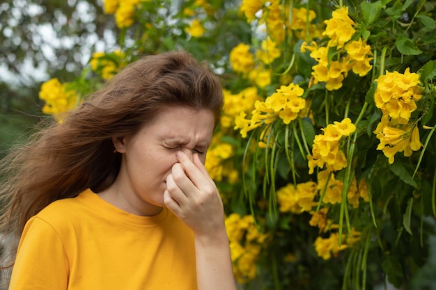 Photo portrait of beautiful young allergic woman is suffering from pollen allergy or cold on natural flower flowering tree background at spring or sunny summer day sneezes blowing her runny nose rubs eyes