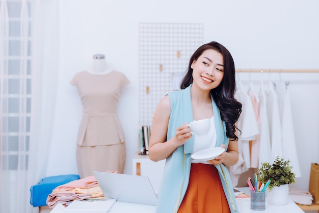 Portrait of beautiful young Asian fashion designer businesswoman at her studio while drinking coffee