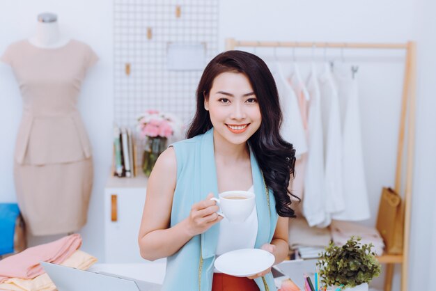 Portrait of beautiful young Asian fashion designer businesswoman at her studio while drinking coffee