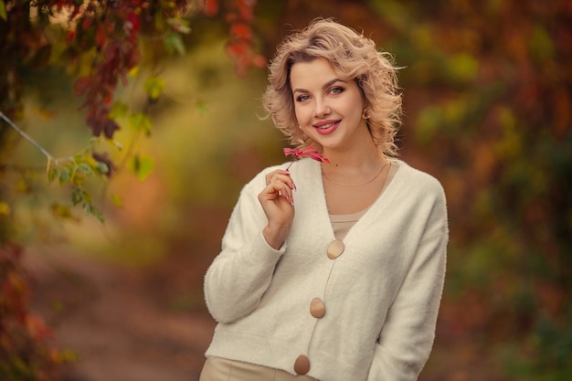 Portrait of a beautiful young blonde woman with a red leaf in her hands in an autumn park.