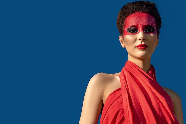 Portrait of a beautiful young brunette woman with creative makeup on her face, who is dressed in a red tunic and stands against a blue hanging sky