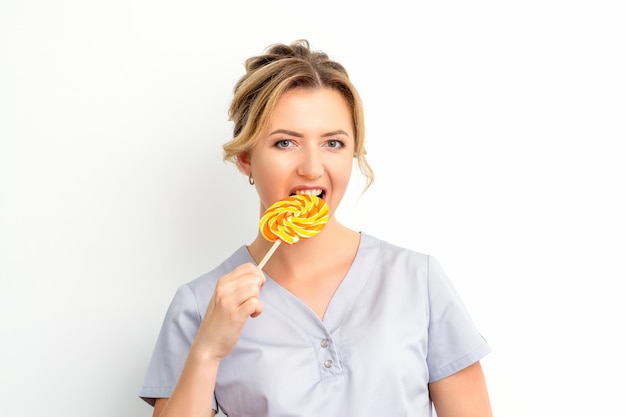 Portrait of a beautiful young caucasian beautician wearing a medical shirt bites a lollipop on a white background