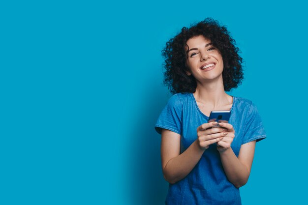 Portrait of a beautiful young curly woman dressed in blue jeans dress laughing while looking at camera isolated on blue studio wall touching her face.