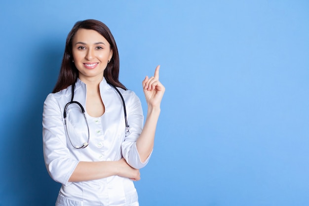 Portrait of a beautiful young doctor of a woman in a white robe on a blue background A place for text and advertising Pediatrician Therapist Home doctor Doctor online