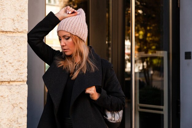 Portrait of a beautiful young model in pink knitted hat standing do to work on background mirrow wall on sunny autumn day in big city Autumn warm photoWoman smiling and look away joyful cheerful mood