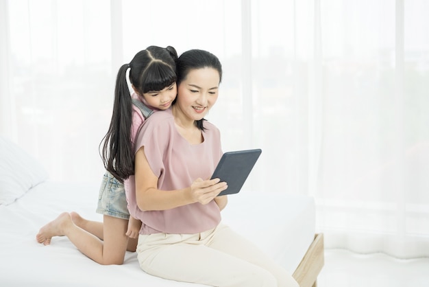 Portrait of beautiful young mom with girl sitting on the bed in a moment of intimacy while buying products online with tablet,
social distancing and online education concept.