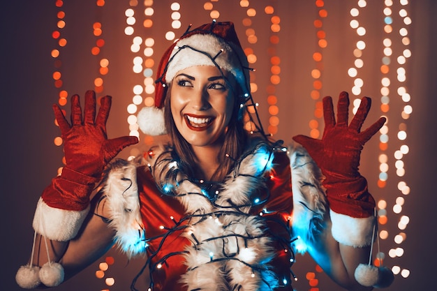 Portrait of a beautiful young smiling woman in Santa Claus costume having fun with christmas light decoration.