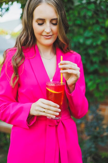 Portrait of a beautiful young woman in a bright pink jacket with a cocktail in her hands Smiling happy woman drinking refreshing lemonade on summer cafe terrace