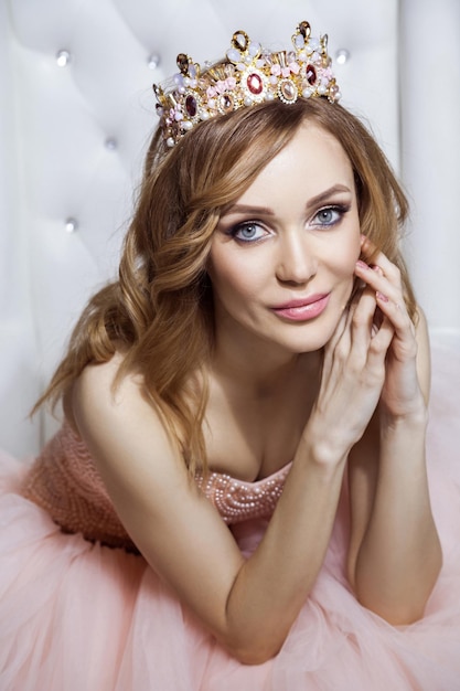 Portrait of beautiful young woman in crown and luxurious pink dress posing on stylish white armchair. sitting, touching her face and looking at camera. indoor studio shot.