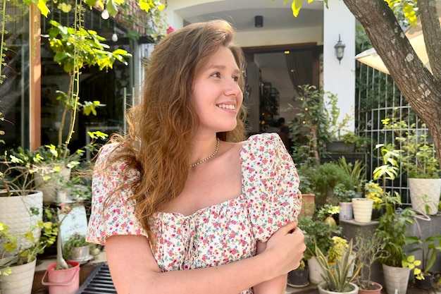 Portrait of beautiful young woman gardener in home fresh natural garden with growing green leaves girl in summer dress is smiling and looking at camera Eco village house or cafe