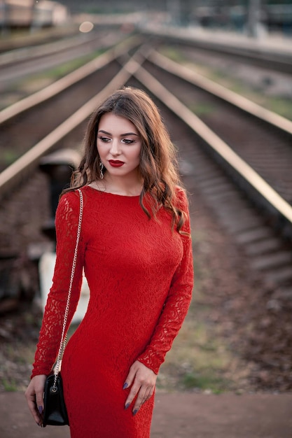 Portrait beautiful young woman in a long red dress stands near the railway