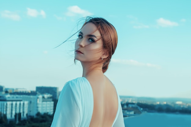 Portrait of beautiful young woman in long white dress standing on rooftop of building with cityscape