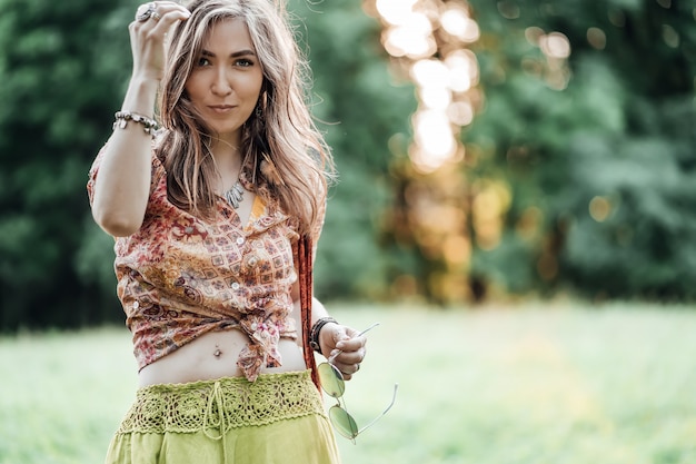 Portrait of Beautiful young woman wearing bohostyle clothes posing in the rays of the evening sun, sunset. Boho style fashion, female wearing silver jewelry having fun in park outdoors.