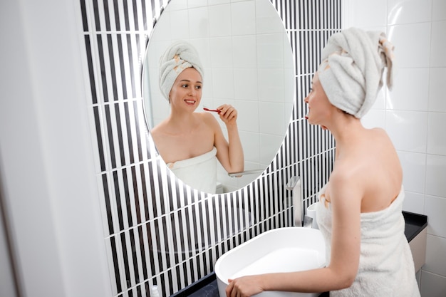 Portrait of a beautiful young woman wearing white towel brushing teeth in front of her bathroom mirror Morning routine beauty procedure