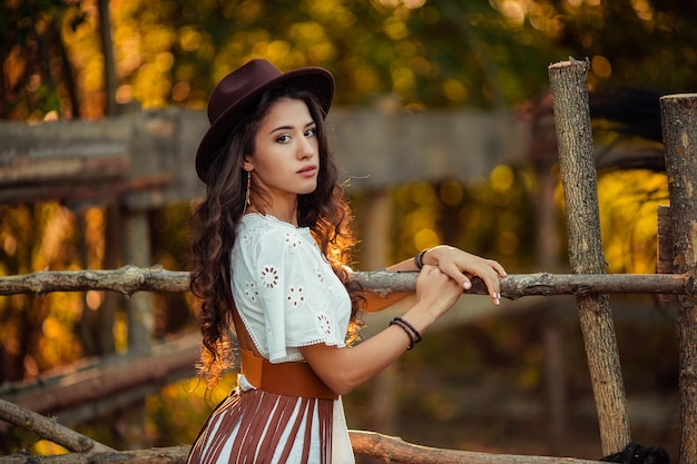 Portrait of a beautiful young woman in a white dress with over the knee boots and a hat with a belt and a fringe on a rustic farm stable