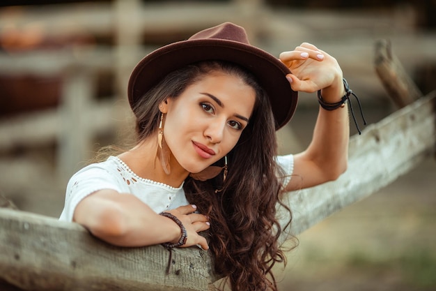 Portrait of a beautiful young woman in a white dress with over the knee boots and a hat with a belt and a fringe on a rustic farm stable