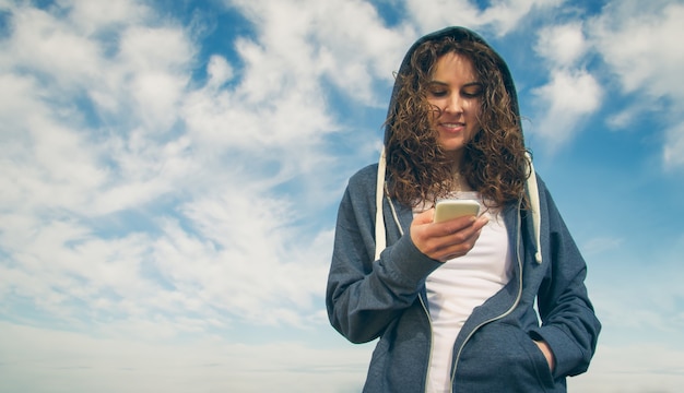 Portrait of beautiful young woman with blue hoodie and sportswear looking her smartphone over a cloudy blue sky background