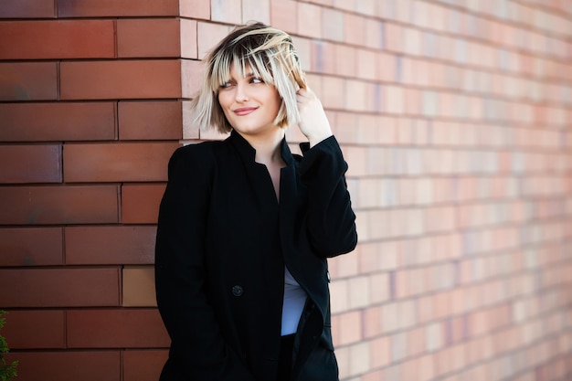 Portrait of a beautiful young woman with short hair in a black coat against a brick wall