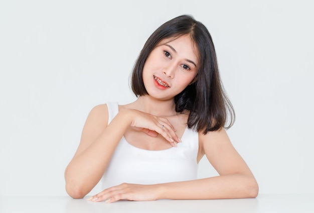 Portrait beauty shot, close up of millennial Asian short black hair model with makeup red lip in tank top undershirt sitting at table posing look at camera on white background.